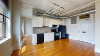 This single-floor loft-style unit blends industrial character with practical living. The open main area is anchored by warm, honey-toned hardwood floors that stretch throughout, complemented by elevated ceilings adorned with exposed silver ductwork and track lighting — hallmarks of a converted urban building. Large double windows frame sweeping city skyline views, flooding the space with natural light and lending an airy, expansive feel.

The kitchen runs along one wall of the open living space and features white cabinetry — including open shelving — paired with black appliances: a refrigerator, electric range, dishwasher, and an overhead range hood. The contrasting palette gives the kitchen a clean, classic look that integrates naturally into the open-plan layout.

A separate bathroom offers a full-size tub and shower combo with white surround, a vanity with dark countertop, and neutral tile flooring. Notably, a stacked washer and dryer unit is conveniently housed within the bathroom, providing in-unit laundry. A louvered closet door near the entry adds additional storage. The overall ambiance of the space is urban and functional, with industrial-chic details balanced by classic architectural trim and sun-drenched interiors.