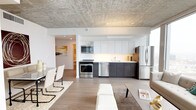 This sophisticated high-rise residence offers a seamless blend of modern industrial design and urban luxury. Exposed concrete ceilings lend an architectural character throughout the space, while light hardwood flooring provides a warm contrast to the raw finishes above.

The open-concept living and dining area is anchored by expansive floor-to-ceiling windows that frame sweeping panoramic city skyline views, flooding the space with natural light. A private balcony extends the living area outdoors, perfect for taking in the urban landscape. Structural concrete columns add to the industrial aesthetic without compromising the sense of openness.

The sleek, galley-style kitchen is outfitted with two-tone cabinetry — gloss white uppers paired with dark lower cabinets — complemented by a clean white countertop and a full suite of stainless steel appliances, including a range, built-in microwave, dishwasher, and refrigerator.

Two well-proportioned bedrooms each feature the signature exposed concrete ceilings and large windows with city views, offering a tranquil retreat above the urban environment. Both bathrooms are finished in a clean, minimalist style with full soaking tubs, tile surrounds, dark vanity cabinetry, and large mirrors with integrated lighting.