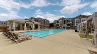 This single-floor residence is part of a well-appointed modern community distinguished by its craftsman-inspired architecture, featuring a combination of grey siding, stone accents, and metal roofing across multi-story buildings. The outdoor amenity area is a standout feature, centered around a large, resort-style swimming pool surrounded by a generous concrete deck and lounge seating — ideal for relaxing under open skies. A covered pavilion with columned supports provides shaded outdoor gathering space adjacent to the pool.

Inside, the interior design blends contemporary and industrial elements with a refined sensibility. The dining area serves as a focal point, anchored by a striking round green marble table paired with woven rattan chairs atop a natural fiber rug. Polished concrete floors run throughout, adding an industrial-chic character that contrasts elegantly with the crisp white walls. A barn-style sliding door adds architectural interest and functional versatility to the space.

Framed wall art enhances the curated, gallery-like atmosphere of the interior. Doorways connect the dining area to additional rooms beyond, where more framed artwork and warm lighting continue the aesthetic. The neutral, light-filled palette throughout creates a calm and sophisticated ambiance, making this residence both visually compelling and functionally comfortable.