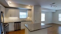 This single-floor space showcases a clean, modern design centered around an open-concept kitchen that flows seamlessly into the adjoining living areas. The kitchen is appointed with crisp white shaker-style cabinetry that extends to the upper walls, providing generous storage. A standout granite island with a bold speckled pattern anchors the space, offering both a functional workspace and a natural gathering point. Matching granite countertops line the perimeter, complementing the stainless steel dishwasher and undermount sink. Recessed lighting supplements the natural light that pours in through multiple windows dressed with classic white horizontal blinds, giving the space a bright and airy feel. A white subway tile backsplash adds a timeless, polished touch to the kitchen walls. Warm hardwood flooring runs throughout, providing a welcoming contrast to the white cabinetry and neutral wall tones. The layout opens toward an adjacent room through a paneled white door, hinting at a connected and functional floor plan. Overall, the space presents a well-appointed, light-filled interior with a cohesive and contemporary aesthetic.