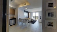 This contemporary single-floor residence showcases a thoughtfully designed open-concept layout that seamlessly connects the kitchen, dining, and living areas into one cohesive living space. The interior is finished with large-format light-toned ceramic tile flooring throughout, contributing to a clean, expansive aesthetic.

The kitchen, positioned to the left of the entryway, features warm wood cabinetry, stainless steel appliances, and efficient use of space. A defined entryway with a recessed ceiling detail creates a natural transition into the dining area, where a sleek white dining table with matching upholstered chairs comfortably accommodates multiple guests.

The living area flows directly from the dining space and is anchored by a dark area rug, a wall-mounted flat-screen television, and a contemporary seating arrangement. Floor-to-ceiling windows with sheer white curtains line the far wall, allowing abundant natural light to fill the space while framing open outdoor views beyond a private balcony. The overall ambiance is modern, bright, and airy, with a neutral white-and-grey palette accented by warm wood tones.
