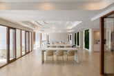 This expansive single-floor space is designed with a refined, contemporary aesthetic that seamlessly blends functionality with upscale design. Light-wash hardwood flooring runs throughout, complementing a crisp white palette and coffered ceiling details accented by recessed lighting. Floor-to-ceiling windows with sheer drapery line multiple areas, flooding the interior with natural light and offering panoramic outdoor views.

The open-plan layout is organized into distinct zones. A central lounge area features generous sectional seating in neutral tones, anchored by area rugs and accent tables, alongside a separate seating zone furnished with sculptural rose-toned velvet chairs. A bar-height communal table with woven rattan stools serves as a casual gathering and working hub.

A row of glass-enclosed private offices or meeting rooms with warm wood-framed doors runs along one wall, providing focused workspace within the open environment. Striking living green walls — lush vertical gardens — serve as bold biophilic accents throughout the space. Additional amenities include a wet bar with bronze cabinetry and what appears to be a golf simulator area. The overall ambiance is polished, airy, and thoughtfully curated for both productivity and leisure.