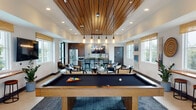 This single-floor amenity lounge is thoughtfully designed to balance recreational activity with comfortable social gathering. The space is defined by its striking slatted wood ceiling, which anchors the central games area and adds warmth to the otherwise bright, modern interior. Recessed lighting and a cluster of pendant lights suspended over the billiards table provide focused illumination, complementing the generous natural light that streams in through multiple windows dressed with woven bamboo shades.

The games area serves as the focal point of the space, featuring a full-size pool table, a foosball table, and a shuffleboard setup. Adjacent to this zone, a casual lounge area offers a leather sofa, rattan-framed armchairs, and a glass-top coffee table arranged around a wall-mounted flat-screen television. A built-in counter with bar stools runs along one wall, providing additional seating with views to the exterior.

Toward one end of the space, a well-appointed kitchenette includes rich walnut cabinetry, stainless steel appliances, a sink with a patterned tile backsplash, and a wall-mounted television. A dining table with chairs nearby creates a dedicated space for group meals or casual gatherings. The overall ambiance is modern and inviting, with layered textures and curated décor throughout.