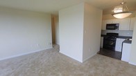 This single-floor residential space offers a clean, functional layout designed for comfortable everyday living. The interior is finished with a neutral, warm color palette throughout, creating a bright and welcoming atmosphere. The main living area features soft carpeting underfoot and is characterized by its open feel, with a partial wall providing a natural visual division between the living space and the adjacent areas. White baseboards run along the perimeter, adding a crisp, polished detail to the rooms. A hallway visible from the main living area connects to additional spaces within the home, hinting at a well-organized floor plan. The kitchen, partially visible to one side, transitions to tile flooring and includes white upper cabinetry along with dark countertops, providing a practical and classic contrast. The overall ambiance of the space is understated and move-in ready, with a straightforward design that prioritizes functionality. The consistent neutral tones across walls and floors lend a cohesive quality to the interior, making it a versatile canvas suitable for a variety of furnishing styles and personal touches.