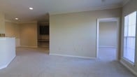 This single-floor space presents a clean, functional layout highlighted by consistent design choices throughout. The main living area is finished with plush, light-colored wall-to-wall carpeting that adds warmth underfoot, while neutral beige walls create a versatile backdrop suited to a variety of décor styles. Crown molding runs along the ceiling perimeter, lending a subtle architectural refinement to the otherwise understated interior.

A doorway on the right side of the main room leads to an adjacent carpeted space, suggesting a bedroom or secondary living area that mirrors the same neutral palette. To the rear left, a built-in workspace is visible, featuring dark cabinetry mounted above a built-in desk or counter surface — a practical and permanent feature well-suited for a home office setup or study nook.

Recessed lighting is installed in the ceiling, providing even ambient illumination throughout the space. White baseboards cleanly frame the flooring along each wall, reinforcing the tidy, well-maintained character of the interior. Overall, the space conveys a calm, move-in-ready atmosphere with functional built-ins and cohesive finishes.