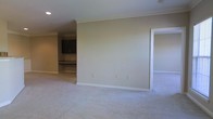 This single-floor space presents a clean, functional layout highlighted by consistent design choices throughout. The main living area is finished with plush, light-colored wall-to-wall carpeting that adds warmth underfoot, while neutral beige walls create a versatile backdrop suited to a variety of décor styles. Crown molding runs along the ceiling perimeter, lending a subtle architectural refinement to the otherwise understated interior.

A doorway on the right side of the main room leads to an adjacent carpeted space, suggesting a bedroom or secondary living area that mirrors the same neutral palette. To the rear left, a built-in workspace is visible, featuring dark cabinetry mounted above a built-in desk or counter surface — a practical and permanent feature well-suited for a home office setup or study nook.

Recessed lighting is installed in the ceiling, providing even ambient illumination throughout the space. White baseboards cleanly frame the flooring along each wall, reinforcing the tidy, well-maintained character of the interior. Overall, the space conveys a calm, move-in-ready atmosphere with functional built-ins and cohesive finishes.