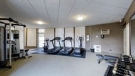 This fitness center offers a practical and functional environment for residents to maintain an active lifestyle. The space is outfitted with a selection of cardio equipment, including two treadmills, a stationary bike, and an elliptical trainer, all positioned on protective rubber matting over neutral carpeted flooring. A drop tile ceiling with flush-mounted lighting provides consistent, even illumination throughout the room. Natural light filters in through vertical blind-covered windows along one wall, adding brightness to the space. An exposed brick accent wall on the opposite side lends a subtle industrial character to the room. A wall-mounted HVAC unit ensures year-round climate comfort. Additional fitness equipment, including a workout bench, is visible along the perimeter, rounding out the available amenities. The overall layout is open and uncluttered, allowing for easy movement between equipment. The clean, no-frills design keeps the focus on functionality, making this fitness center a convenient and accessible option for everyday workouts.