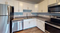 This single-floor unit offers a thoughtfully designed layout with a clean, modern aesthetic throughout. The open living area features durable wood-look luxury vinyl flooring in a warm tone, neutral gray walls, and a glass-paneled exterior door that fills the space with natural light and provides outdoor access. The kitchen is well-equipped with crisp white shaker-style cabinetry, granite countertops, stainless steel appliances including a dishwasher and range, and a double-basin sink — combining both style and function. Multiple bedrooms are finished with plush gray carpet, ceiling fans with light fixtures, and baseboard heating. The larger bedroom benefits from corner window placement, allowing abundant natural light and outdoor views from multiple angles, while additional bedrooms each feature their own windows. The bathrooms are finished with wood-look vinyl flooring, white vanity cabinetry, granite countertops, and brushed nickel hardware. A large mirror enhances the sense of space in the full bath. Throughout the unit, consistent neutral tones, white six-panel doors, and clean baseboards contribute to a cohesive and contemporary feel.