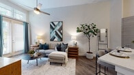 This single-floor residence showcases a thoughtfully designed open layout with a consistent modern aesthetic throughout. The living area is highlighted by notably tall ceilings and a ceiling fan, creating an airy atmosphere. Large windows dressed with blinds and teal drapes allow generous natural light to fill the space, complementing the warm wood-look flooring found throughout. The room comfortably accommodates a sectional sofa and a round coffee table, providing a welcoming and functional gathering space.

The kitchen is outfitted with sleek, flat-front cabinetry in a warm gray tone, a white quartz countertop, a full suite of stainless steel appliances including a French door refrigerator, built-in microwave, and dishwasher. A prominent kitchen island with pendant lighting adds both prep space and a casual dining option. Under-cabinet lighting and a linear tile backsplash contribute a refined, contemporary feel.

The bedroom features tall ceilings, natural window light, and direct access to both a dedicated walk-in closet and a private en-suite bathroom. The bathroom is finished with quartz countertops, a glass-enclosed walk-in shower, wood-look tile flooring, and neutral warm-toned walls, completing the cohesive, modern character of the space.