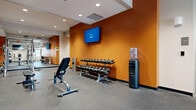 This fitness center is a comprehensively outfitted facility designed to accommodate a wide variety of exercise needs. The space is organized across a single open floor, with distinct zones dedicated to different training disciplines. Bold orange accent walls punctuate the interior, lending the gym an energetic and motivating atmosphere, while recessed ceiling lighting ensures consistent, even illumination throughout.

The free weights area features a full dumbbell rack with a wide range of weights, an adjustable bench, and a wall-mounted flat-screen television for entertainment during workouts. A water cooler station is conveniently positioned nearby. Adjacent to this zone, a strength training section houses multiple cable and resistance machines, thoughtfully arranged to allow easy access and safe use.

Floor-to-ceiling mirrors line one wall, visually expanding the space and allowing users to monitor their form during lifts and exercises. The cardio zone is equipped with treadmills, ellipticals, upright bikes, and additional cardio machines, complemented by wall-mounted televisions. A dedicated powerlifting area includes a full Smith machine rack with weight plates and a rowing machine. The durable rubber flooring throughout provides excellent traction and impact absorption, making this fitness center a functional and well-appointed training environment.