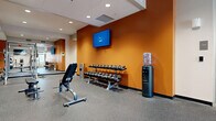 This fitness center is a comprehensively outfitted facility designed to accommodate a wide variety of exercise needs. The space is organized across a single open floor, with distinct zones dedicated to different training disciplines. Bold orange accent walls punctuate the interior, lending the gym an energetic and motivating atmosphere, while recessed ceiling lighting ensures consistent, even illumination throughout.

The free weights area features a full dumbbell rack with a wide range of weights, an adjustable bench, and a wall-mounted flat-screen television for entertainment during workouts. A water cooler station is conveniently positioned nearby. Adjacent to this zone, a strength training section houses multiple cable and resistance machines, thoughtfully arranged to allow easy access and safe use.

Floor-to-ceiling mirrors line one wall, visually expanding the space and allowing users to monitor their form during lifts and exercises. The cardio zone is equipped with treadmills, ellipticals, upright bikes, and additional cardio machines, complemented by wall-mounted televisions. A dedicated powerlifting area includes a full Smith machine rack with weight plates and a rowing machine. The durable rubber flooring throughout provides excellent traction and impact absorption, making this fitness center a functional and well-appointed training environment.