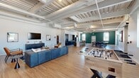 This generously sized, single-floor amenity space offers a thoughtfully designed open-concept layout that seamlessly blends recreation, socializing, and productivity. The interior is defined by soaring ceilings with exposed structural decking and visible ductwork, lending an industrial-modern aesthetic throughout. Light hardwood flooring flows continuously across all areas, unifying the space.

The main lounge area features a large sectional sofa, accent chairs, and a wall-mounted television, creating a comfortable gathering spot. Adjacent to the lounge, a foosball table anchors the recreation zone, which benefits from abundant natural light through oversized black-framed windows. A bold accent wall with multicolored horizontal stripes in burgundy, gold, gray, and teal adds a vibrant focal point and leads to a set of glass-paned French doors opening to a separate office or meeting room.

The full kitchen is well-appointed with dark cabinetry, granite countertops, white subway tile backsplash, stainless steel appliances including a refrigerator and built-in microwave, and a double sink. A dining area with bar seating and tables is positioned near a teal feature wall and windows overlooking the exterior. The space is bright, functional, and designed to accommodate a variety of communal activities.