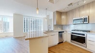 Located in the heart of downtown Phoenix, this thoughtfully designed 1-bedroom, 2-bathroom residence offers generous living space across a single floor. Warm hardwood-style flooring flows throughout, complemented by crisp white walls and abundant natural light pouring in through expansive floor-to-ceiling windows that frame sweeping views of the Phoenix skyline.

The open-concept kitchen and living area serve as the centerpiece of the residence. The contemporary kitchen features two-tone cabinetry — white lower cabinets paired with natural wood upper cabinets — marble-style tile backsplash, stone countertops, a central island with bar seating, stainless steel appliances including a gas range, dishwasher, microwave, and refrigerator, plus stylish pendant lighting. The living room is generously proportioned with panoramic city views on multiple sides.

The bedroom is a bright, serene retreat with corner floor-to-ceiling windows offering commanding skyline vistas. Two closets provide ample storage, including a walk-in with wire shelving. The primary bathroom is spa-inspired, featuring a dual-sink vanity, a soaking tub, and a separate glass-enclosed shower with tile surround. A second bathroom includes a glass shower and in-unit stacked washer and dryer for added convenience.