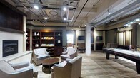 This single-floor communal lounge space blends industrial and contemporary design elements to create a sophisticated and inviting environment. The open-plan layout is defined by exposed ceiling ductwork, structural concrete columns, and recessed track lighting, lending the space a modern, loft-inspired aesthetic. The flooring features a combination of wood-look planks and patterned area rugs that help delineate distinct activity zones throughout the room. A generous seating area is anchored by plush upholstered armchairs arranged around a round wooden coffee table, ideal for relaxed conversation or casual gatherings. A built-in dark wood bookcase lines one wall, displaying books and decorative objects, adding warmth and a library-like character to the space. Multiple flat-screen televisions are mounted throughout the room, ensuring excellent visibility from various vantage points. Towards the rear of the space, a billiards table provides an entertainment focal point, complemented by additional seating and bar-height tables. Natural light filters in through windows, softening the industrial elements and contributing to an overall atmosphere that is both energetic and comfortable — well-suited for socializing, recreation, and leisure.