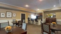 This single-floor space exudes a warm and inviting atmosphere, thoughtfully designed to accommodate both casual gatherings and more formal occasions. The interior is finished with rich, warm yellow walls accented by classic white crown molding, lending an air of traditional elegance throughout. A textured ceiling with recessed lighting provides a polished overhead detail that ties the space together.

The dining area is furnished with dark wood tables and upholstered chairs in a muted blue-green patterned fabric, offering a refined yet comfortable setting for meals and socializing. A decorative floral centerpiece adds a welcoming touch to the dining tables. Framed artwork adorns the walls, contributing to the space's curated, homey character.

A kitchenette is seamlessly integrated into the space, featuring dark wood cabinetry, dark countertops, a refrigerator, and a microwave. Bar-height seating lines the counter, making it a practical and social hub. A window above the counter allows natural light to brighten the kitchen area.

Plush, neutral-toned carpeting throughout adds warmth underfoot, while the open layout ensures ease of movement. A dark-painted door provides access to an adjacent area, completing this well-appointed, multifunctional community space.