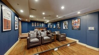 This single-floor movie lounge is thoughtfully designed to deliver an authentic cinematic experience. The space is defined by its bold navy blue walls, which set a dramatic and immersive tone throughout. Recessed lighting overhead provides warm, even illumination that complements the theater-inspired ambiance.

The seating area features plush, dark brown leather club chairs arranged in a tiered layout, ensuring clear sightlines to the large flat-screen display mounted on the front wall. A built-in media console with warm wood cabinetry and a light countertop houses the audiovisual equipment. In-ceiling speakers are positioned throughout the room to support a full surround-sound experience.

A curated gallery of framed movie posters lines the walls, showcasing a wide range of cinematic classics and blockbusters — from Back to the Future and The Lion King to Marvel titles such as Infinity War, Endgame, and Black Panther — adding a lively, pop-culture energy to the décor. Round wood-and-glass side tables are positioned between seating clusters for added convenience.

A countertop bar area with bar-height seating offers a space for refreshments, enhancing the social aspect of the lounge. Warm-toned drape curtains on one wall add a classic theater touch. The patterned carpet flooring ties together the rich color palette, making this space an inviting destination for film enthusiasts.