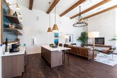 This thoughtfully designed single-floor communal space blends contemporary and mid-century modern aesthetics to create a warm, inviting atmosphere. The open-plan layout is anchored by soaring ceilings adorned with exposed wood beams, lending a rustic yet refined character throughout. Rich, dark hardwood flooring runs the length of the space, providing a grounding contrast to the crisp white walls.

A dramatic floor-to-ceiling stacked-stone fireplace serves as the focal point of the lounge area, which is furnished with a leather Barcelona-style chair, tufted seating, and layered area rugs. Multiple large windows flood the interior with natural light and offer views of the surrounding exterior. Wall-mounted sconces complement the statement chandelier above, creating a well-layered lighting scheme.

A dedicated dining and seating section features teal upholstered banquette seating paired with round marble-top tables and woven accent chairs — ideal for casual gatherings or collaborative use. Adjacent to this area is a well-appointed kitchenette, highlighted by a bold teal herringbone tile backsplash, light wood cabinetry, quartz countertops, open floating shelves, brass fixtures, and a central island. Stainless steel appliances complete the functional setup. Together, these zones create a versatile, design-forward space suited for socializing, lounging, and everyday use.