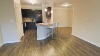 This single-floor space showcases an open-concept layout that seamlessly connects the kitchen and living areas, creating a cohesive and functional environment. The kitchen is appointed with rich, dark espresso cabinetry that extends from floor to ceiling, providing ample storage. A classic tile backsplash complements the cabinetry, while black appliances reinforce the bold, contrast-driven aesthetic. The centerpiece of the kitchen is a curved peninsula topped with granite countertops featuring warm, earthy tones. Two contemporary white barstools with chrome legs are positioned at the peninsula, creating a casual dining or entertaining space. Pendant lighting hangs above the peninsula, adding focused illumination and visual interest. Wide-plank, wood-tone flooring in a rustic brown finish runs throughout both the kitchen and adjacent living area, grounding the space with warmth and continuity. Crown molding traces the perimeter of the ceiling, lending an elevated, finished look. The open living area beyond the kitchen offers generous room for furniture arrangement, with neutral warm beige walls providing a versatile backdrop. Track lighting in the kitchen ensures the workspace is well-lit throughout the day.