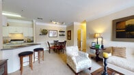 Located at 3920 Club Dr in Duluth, Georgia, this single-floor residence offers a thoughtfully designed layout that balances comfort and functionality. The entryway opens to a spacious living room finished with plush carpeting, crown molding, and generous natural light streaming through large windows dressed with decorative curtains. The living area flows seamlessly into a dedicated dining space, which features wall-mounted shelving and a glass-top dining table — ideal for everyday meals or entertaining guests. A breakfast bar with bar stools connects the dining area to the kitchen, which is outfitted with white cabinetry, dark countertops, a double sink, dishwasher, and full-size refrigerator, all set atop warm hardwood flooring. The primary bedroom is a serene retreat with carpeted floors, a walk-in closet, and an en suite bathroom featuring tile flooring, a single vanity, and a shower-tub combination. The second bedroom is equally comfortable, offering carpeted floors, a large window, and convenient access to a second full bathroom with a similar layout. Warm, neutral tones and crown molding details throughout give this residence a cohesive and welcoming ambiance.