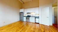 Located at 145 Clifford St in Providence, Rhode Island, this single-floor unit showcases an open, loft-inspired layout defined by impressively tall ceilings and warm, honey-toned hardwood floors that run throughout the living and bedroom areas. The generous ceiling height lends an airy, expansive quality to the space, enhanced further by large arched windows that invite natural light deep into the interior.

The kitchen is well-appointed with a full suite of stainless steel appliances, including a refrigerator, electric range, dishwasher, and built-in microwave venthood. Crisp white shaker-style cabinetry extends from counter height to the upper walls, maximizing storage, while dark stone countertops provide a clean contrast. The layout allows for an efficient workflow and connects naturally to the open living area.

The bedroom is a calm, understated space with the same rich hardwood flooring and clean white walls, offering a versatile canvas. The bathroom is finished with classic white subway tile surrounding a full soaking tub and shower, a white vanity with under-sink storage, and light tile flooring, creating a bright and functional space. Overall, the unit carries a modern yet warm character rooted in its loft-style architectural bones.