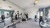 Located at 505 W Baseline Rd in Tempe, Arizona, this fitness center at Harbor at Baseline offers a thoughtfully designed single-floor workout space that blends functionality with aesthetic appeal. The gym features a vaulted ceiling that lends an open, airy feel, complemented by recessed lighting and stylish pendant light fixtures throughout. A striking warm-toned wood plank accent wall serves as a focal point, adding a natural, inviting contrast to the cool teal-painted walls and crisp white ceiling. Floor-to-ceiling mirrors line one side of the space, visually expanding the room and supporting proper form during workouts. Natural light streams in through multiple windows, creating a bright and energizing environment. The fitness center is comprehensively equipped with a wide variety of cardio machines, including treadmills, elliptical trainers, stationary bikes, a rowing machine, and a stair climber. Strength training needs are met with a multi-station cable machine, adjustable weight bench, and a rack of medicine balls. A wall-mounted television provides entertainment during workouts. A set of teal-framed French doors opens to the outdoor area, allowing for easy access and additional ventilation. The space is climate-controlled with a wall-mounted AC unit, ensuring year-round comfort for users.