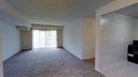 This single-floor residential space offers a clean, functional layout designed for comfortable everyday living. The main living area is a standout feature, characterized by its generous proportions, neutral white walls, and plush wall-to-wall carpeting that adds warmth underfoot. A large sliding glass door floods the room with natural light and provides direct access to an outdoor area, while full-length vertical blinds offer privacy and light control as needed. The open layout of the living space allows for flexible furniture arrangements, making it easy to customize to personal taste. A partial dividing wall creates a subtle architectural distinction between areas, hinting at a separate adjoining space — likely a dining area or entryway — without fully closing off the floor plan. The overall color palette is understated and neutral, featuring soft whites and warm grays that contribute to a clean, move-in-ready aesthetic. The space is well-suited for those seeking a straightforward, low-maintenance residence with ample natural light and an open, airy feel throughout.