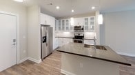 This single-floor space showcases a cohesive modern design with neutral tones and quality finishes throughout. The kitchen serves as a central highlight, outfitted with crisp white shaker-style cabinetry — including glass-front upper cabinets — paired with sleek dark countertops. Stainless steel appliances, including a French door refrigerator with ice dispenser, electric range, and over-the-range microwave, provide a polished, functional workspace. A substantial island with an undermount sink and pendant lighting above adds both prep space and visual appeal. Light wood-look flooring flows throughout the open-concept kitchen and living areas, complementing the bright, clean aesthetic.

The bedroom features soft carpeting underfoot, neutral gray walls, and a ceiling fan with integrated lighting. A horizontal clerestory window allows natural light while maintaining privacy. The bathroom is neatly appointed with a white vanity cabinet, gray stone-look countertop, undermount sink, a modern chrome faucet, and a large framed mirror. Consistent use of neutral palettes, recessed lighting, and clean architectural lines gives the space a fresh, move-in-ready feel throughout.