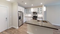 This single-floor space showcases a cohesive modern design with neutral tones and quality finishes throughout. The kitchen serves as a central highlight, outfitted with crisp white shaker-style cabinetry — including glass-front upper cabinets — paired with sleek dark countertops. Stainless steel appliances, including a French door refrigerator with ice dispenser, electric range, and over-the-range microwave, provide a polished, functional workspace. A substantial island with an undermount sink and pendant lighting above adds both prep space and visual appeal. Light wood-look flooring flows throughout the open-concept kitchen and living areas, complementing the bright, clean aesthetic.

The bedroom features soft carpeting underfoot, neutral gray walls, and a ceiling fan with integrated lighting. A horizontal clerestory window allows natural light while maintaining privacy. The bathroom is neatly appointed with a white vanity cabinet, gray stone-look countertop, undermount sink, a modern chrome faucet, and a large framed mirror. Consistent use of neutral palettes, recessed lighting, and clean architectural lines gives the space a fresh, move-in-ready feel throughout.