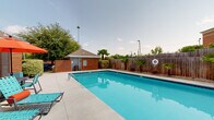 This outdoor pool area is a well-designed recreational space centered around a generously sized rectangular swimming pool with clear, bright turquoise water. The pool is finished with classic tiled edges and includes entry steps for easy access, with a safety ring mounted on the surrounding privacy fence. The concrete pool deck provides ample space for lounging, furnished with multiple teal-framed chaise lounges and patio seating accented by orange umbrellas, creating a vibrant and inviting atmosphere.

The pool area is enclosed by a tall wooden privacy fence, which is thoughtfully landscaped with small palm trees and low shrubs along its base. A brick pool house with a pitched shingle roof anchors one end of the space, featuring a covered porch with seating, large windows, and posted pool rules — suggesting it serves as both a functional utility building and a social gathering spot. Additional mature trees frame the perimeter, providing shade and a natural backdrop. The overall setting is open to the sky, allowing for abundant natural light throughout the day, making this a well-equipped and attractive outdoor amenity.
