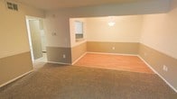 This single-floor space offers an open, connected layout that integrates the living, dining, and kitchen areas into a cohesive flow. The living area is finished with wall-to-wall carpet in a warm beige tone, creating a comfortable and inviting atmosphere. Adjacent to it, the dining area transitions to smooth laminate wood flooring, providing a practical and visually distinct zone for dining. The walls feature a two-tone paint scheme, with a darker gray-taupe on the lower half and a lighter neutral tone above, adding subtle visual interest throughout the space. A pass-through window connects the dining area to the kitchen, enhancing the open feel while maintaining defined spaces. A pendant light fixture hangs above the dining area, serving as a functional and decorative focal point. The kitchen, partially visible through the pass-through and a connecting hallway, features white cabinetry. White baseboards and trim provide clean, crisp contrast against the wall colors. The overall ambiance is calm and neutral, offering a blank canvas well-suited for a variety of interior styles and furnishing arrangements.