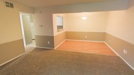 This single-floor space offers an open, connected layout that integrates the living, dining, and kitchen areas into a cohesive flow. The living area is finished with wall-to-wall carpet in a warm beige tone, creating a comfortable and inviting atmosphere. Adjacent to it, the dining area transitions to smooth laminate wood flooring, providing a practical and visually distinct zone for dining. The walls feature a two-tone paint scheme, with a darker gray-taupe on the lower half and a lighter neutral tone above, adding subtle visual interest throughout the space. A pass-through window connects the dining area to the kitchen, enhancing the open feel while maintaining defined spaces. A pendant light fixture hangs above the dining area, serving as a functional and decorative focal point. The kitchen, partially visible through the pass-through and a connecting hallway, features white cabinetry. White baseboards and trim provide clean, crisp contrast against the wall colors. The overall ambiance is calm and neutral, offering a blank canvas well-suited for a variety of interior styles and furnishing arrangements.