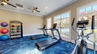 This fitness center at 8064 S Fulton Pkwy in South Fulton, Georgia, offers a well-equipped, single-floor exercise space designed to support a well-rounded workout routine. The room is finished with warm neutral-toned walls, crisp white crown molding, and bold blue patterned carpet flooring that adds a modern, energetic aesthetic throughout.

Large multi-pane windows line the walls, flooding the space with abundant natural light and providing views of the outdoor pool area and surrounding tree-lined landscape. Dual ceiling fans with integrated lighting ensure comfortable airflow during workouts.

The cardio section is outfitted with multiple TRUE brand treadmills and an elliptical trainer, all positioned to take advantage of the pool-facing windows. A dedicated strength training area features a two-tier dumbbell rack with a range of free weights, a flat workout bench, and a medicine ball storage rack stocked with an assortment of weighted balls. Wall-mounted flat-screen televisions provide entertainment while exercising.

Recessed lighting supplements the natural light, maintaining a bright and inviting atmosphere throughout the day. The space is thoughtfully arranged to allow ample room for movement and stretching, with foam rollers also available. The overall ambiance is clean, functional, and energizing, making it a practical and appealing fitness amenity.