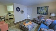This single-floor residence offers a warm and inviting atmosphere, highlighted by an eclectic and colorful interior design sensibility. The living area serves as the heart of the space, featuring rich hardwood-style flooring that grounds the room with a sense of warmth. A textured ceiling adds architectural character overhead, while the white walls provide a clean, neutral backdrop that allows the bold furnishings and décor to take center stage. The living room is furnished with vibrant, paisley-patterned seating and an oversized upholstered ottoman that doubles as a coffee table, all resting atop a decorative area rug. A large, colorful abstract painting adorns one wall, while eclectic wall décor, including decorative circular pieces, adds a personal and artistic touch. A vase of white orchids on the ottoman centerpiece brings a touch of nature indoors. A doorway visible from the living area hints at additional rooms, and a glimpse into an adjacent kitchen area suggests a connected, open flow between the living and culinary spaces. Overall, the space exudes a lived-in, expressive charm with a cozy and personalized ambiance throughout.