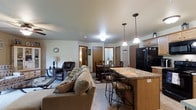 This single-floor residence is designed around an open-concept layout that integrates the kitchen, dining, and living areas into one cohesive, flowing space. The kitchen features a granite-topped breakfast bar with seating for casual dining, pendant lighting overhead, light wood cabinetry, and black appliances, including a full-size refrigerator. The bar counter serves as a natural boundary between the kitchen and the rest of the living space without disrupting the open feel.

Adjacent to the kitchen, a dedicated dining area accommodates a full table and chair set, lit by a second pendant light that defines the zone within the open floor plan. Multiple doorways visible from the main area lead to additional rooms, suggesting a well-connected layout throughout the unit.

The living area is generously proportioned, furnished with a plush sofa, accent pillows, and a leather recliner, creating a comfortable and inviting atmosphere. Neutral-toned carpet and light-colored walls contribute to a bright, airy feel. Recessed and flush-mount ceiling lighting provides consistent illumination throughout. The overall ambiance is warm, practical, and well-suited for everyday living.