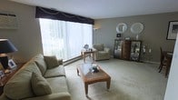 This single-floor space offers a well-appointed and inviting living environment designed with comfort and style in mind. The living room serves as the focal point of the space, immediately drawing attention with its expansive floor-to-ceiling windows dressed with vertical blinds and a draped navy valance, allowing an abundance of natural light to fill the room. The soft, neutral-toned carpeting underfoot adds warmth and comfort throughout. The living room is furnished with a generous sofa and a coordinating armchair, both upholstered in a muted beige fabric, arranged around a wooden coffee table featuring glass panel insets. A tall arc floor lamp provides additional ambient lighting, complementing the natural brightness of the room. Along the wall, a dark wood display cabinet with glass doors showcases decorative collectibles and adds a refined touch to the space. Decorative circular mirrors mounted on the wall further enhance the room's elegant aesthetic. The neutral gray wall tones create a calm, cohesive backdrop that ties the furnishings and décor together seamlessly. A baseboard heating unit beneath the windows reflects the practical infrastructure of the space. Overall, the living area exudes a relaxed yet polished ambiance, making it a welcoming and functional environment.