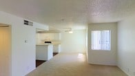 This single-floor space features an open-concept layout that connects the kitchen and living area into one cohesive, functional space. The kitchen is outfitted with white cabinetry, a peninsula-style counter, and white appliances, including a range and dishwasher. Tile flooring in the kitchen transitions smoothly into carpet throughout the living area, adding warmth underfoot. A ceiling fan is centrally positioned overhead, providing both lighting and air circulation. The textured popcorn ceiling runs throughout, adding a classic residential character. A large window fitted with vertical blinds allows abundant natural light to fill the space, creating a bright and airy atmosphere. The neutral white walls and light tones throughout contribute to an open, clean aesthetic. The open floor plan allows for flexible furniture arrangement, making the living area adaptable to various lifestyle needs. Overall, the space presents a straightforward, functional design with practical finishes suited for comfortable everyday living.