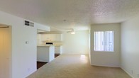 This single-floor space features an open-concept layout that connects the kitchen and living area into one cohesive, functional space. The kitchen is outfitted with white cabinetry, a peninsula-style counter, and white appliances, including a range and dishwasher. Tile flooring in the kitchen transitions smoothly into carpet throughout the living area, adding warmth underfoot. A ceiling fan is centrally positioned overhead, providing both lighting and air circulation. The textured popcorn ceiling runs throughout, adding a classic residential character. A large window fitted with vertical blinds allows abundant natural light to fill the space, creating a bright and airy atmosphere. The neutral white walls and light tones throughout contribute to an open, clean aesthetic. The open floor plan allows for flexible furniture arrangement, making the living area adaptable to various lifestyle needs. Overall, the space presents a straightforward, functional design with practical finishes suited for comfortable everyday living.