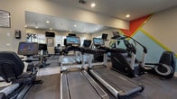 This fitness center is a single-story amenity space designed to support a well-rounded exercise routine. The interior is bright and functional, featuring recessed lighting, smooth neutral-toned walls, and large wall-mounted mirrors that visually expand the space and assist with form during workouts. A bold geometric accent wall in red, orange, and yellow adds a dynamic, energizing touch to the cardio area.

The facility is equipped with a row of Life Fitness treadmills, each featuring integrated touchscreen displays, as well as upright stationary bikes with digital consoles. Additional equipment includes elliptical-style machines and a selection of medicine balls, foam rollers, and resistance accessories neatly organized on a dedicated storage rack. A wall-mounted television and clock complete the space.

Natural light filters in through windows overlooking the exterior, creating a connection to the outdoors. A glass door leads directly to the pool area, offering seamless access to the outdoor amenities.

The exterior features a generously sized swimming pool and adjacent spa, surrounded by a broad concrete deck furnished with lounge chairs and shaded umbrella seating. The pool area is framed by mature trees and overlooks the surrounding buildings, providing a resort-like atmosphere ideal for relaxation after a workout.