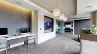 The E-Lounge is a thoughtfully designed single-floor space that seamlessly blends functionality with contemporary style. The interior is anchored by a refined aesthetic, featuring neutral-toned carpet flooring that runs throughout and contributes to the space's calm, professional ambiance.

A standout feature is the striking lighting installation — a cluster of oversized spherical woven pendant lights suspended from the ceiling, casting a warm, diffused glow across the open corridor. The ceiling itself incorporates subtle recessed lighting with warm accent illumination, adding depth and dimension to the space.

A dedicated workstation area is positioned along one wall, complete with a sleek white desk, a task chair with a patterned upholstered seat, and a mounted monitor — ideal for individual productivity. Adjacent to this area, a wall niche framed in warm wood houses a flat-screen display, serving as a central media focal point.

Toward the rear, glass-walled enclosures provide transparency while maintaining acoustic separation, suggesting spaces suited for meetings or focused work. A second display is visible beyond the glass partition, reinforcing the space's tech-integrated design. Textured accent wallcovering adds visual interest and sophistication, completing the polished, modern character of the E-Lounge.