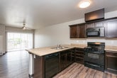 This single-floor unit is organized around an open-concept kitchen and living area, creating a functional and connected layout. The kitchen is well-appointed with dark espresso cabinetry offering ample storage, light granite-style countertops, and black appliances including an electric range, over-the-range microwave, and a double-basin sink set into a peninsula that separates the kitchen from the living space. Wood-look plank flooring runs throughout the main living area, adding warmth and continuity. A ceiling fan is centrally located, and a sliding glass door at the far end of the living space provides access to an outdoor area while allowing natural light to fill the room. The bedroom features neutral carpet, vertical blinds on a window with outdoor views, and baseboard heating for climate control. The bathroom is clean and minimal, finished with wood-look vinyl flooring, a standard toilet, a bathtub with a surrounding surround, and baseboard heating. White walls throughout the unit maintain a neutral, versatile backdrop. The 3D floor plan overview confirms a compact but efficiently arranged single-level footprint with clearly defined zones for living, cooking, sleeping, and bathing.
