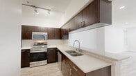 This thoughtfully designed single-floor residence offers a functional and modern living experience across a comfortable footprint. The open-concept kitchen and living area serves as the heart of the home, where the kitchen stands out with rich dark-wood cabinetry, stainless steel appliances including a range and over-the-range microwave, light quartz countertops, a double undermount sink, and track lighting overhead. A pass-through window connects the kitchen to the living area, creating an airy, connected feel. Light wood-look flooring runs consistently throughout the space, complementing the crisp white walls and clean lines found in every room. The spacious primary bedroom features multiple windows that invite abundant natural light and include views of greenery outside, along with a ceiling fan and recessed lighting. An additional den provides flexibility for a home office or guest space. The bathroom is well-equipped with a soaking tub and tiled surround, a dark-wood vanity with light countertop, and brushed nickel fixtures. A dedicated walk-in closet with wire shelving provides generous storage. Completing the layout is an in-unit laundry area, adding convenience to daily living. The overall ambiance is bright, clean, and contemporary.