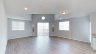 This single-floor space offers an open, airy layout highlighted by elevated ceilings that contribute to a sense of generous volume. The main living area is finished with smooth hardwood-style flooring in a warm neutral tone, lending a clean and contemporary feel underfoot. Soft blue-gray walls create a calm, modern aesthetic throughout the space.

A distinctive circular window positioned near the ceiling serves as a standout architectural detail, drawing the eye upward and introducing an additional source of natural light. Large sliding glass doors at the center of the main wall open to an outdoor area, flooding the interior with daylight and providing a seamless connection to the exterior. Flanking the sliding doors are two symmetrical arched wall niches, adding subtle architectural character and offering decorative display space.

Additional windows fitted with white horizontal blinds line the walls, ensuring the space remains bright and well-lit throughout the day. Recessed and flush-mount ceiling lighting provides practical illumination for evening use. The overall ambiance is clean, open, and inviting, with a neutral palette and thoughtful design details that create a versatile and comfortable living environment.