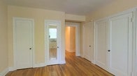 This single-floor space presents a warm and understated aesthetic, characterized by rich hardwood flooring that runs throughout the interior. The walls are finished in a soft, creamy neutral tone that reflects light naturally and contributes to an open, airy feel. Crisp white baseboards and door trim provide clean contrast against the warmer wall tones, adding a classic architectural detail.

The space features multiple doorways, including a distinctive mirrored door that introduces a functional decorative element while visually expanding the room. A central open passage leads to an adjoining room, which is warmly lit and finished in a similarly neutral palette, suggesting a cohesive design throughout. A third solid white-paneled door offers access to an additional area of the space.

Natural light filters in through a window visible in the background, adding brightness and depth to the interior. The overall layout appears well-connected, with rooms flowing naturally from one to the next. The combination of hardwood floors, neutral tones, and white architectural trim gives the space a timeless, clean character that is both practical and visually appealing.