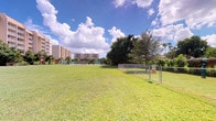 This outdoor community space offers a generous, open-air environment characterized by expansive, well-kept green lawns that invite a wide range of recreational activities. The grounds are framed by mature shade trees, including towering palm trees that lend a distinctly tropical ambiance to the setting. A chain-link fenced area provides a designated zone within the property, complemented by tennis courts visible in the background, making the space well-suited for both leisurely and active use. Picnic tables are conveniently positioned throughout the lawn, offering inviting spots for outdoor gatherings and relaxation beneath the open sky. The surrounding landscape is enriched by lush tropical vegetation, including palms and broadleaf trees that provide natural shade and visual interest. Multi-story residential buildings border the grounds, contributing to a vibrant, community-oriented atmosphere. The space benefits from abundant natural light and open sightlines under a bright, partly cloudy sky, enhancing the overall sense of openness. The well-maintained condition of the grounds, combined with the variety of amenities, reflects a community-focused design intended to support an active and enjoyable outdoor lifestyle in a warm, sunny climate.
