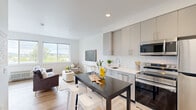Located at 7460 SW Hermoso Way in Tigard, Oregon, this thoughtfully designed studio presents a bright, modern living experience within a efficient single-floor layout. The open-concept kitchen and living area maximizes every inch of the space, creating a seamless flow between cooking, dining, and relaxing zones. The kitchen is outfitted with contemporary gray flat-front cabinetry, white quartz countertops, a white subway tile backsplash, and a full suite of stainless steel appliances including a refrigerator, electric range, over-the-range microwave, and dishwasher. A compact dining table with bar stools serves as a natural divider between the kitchen and living area. The living space is anchored by light hardwood-style flooring and large windows that flood the room with natural light while framing picturesque views of the surrounding Tigard landscape and treetops. The bathroom features a generous single-sink vanity with light blue cabinetry, white countertops, and a large mirror. An in-unit washer and dryer are also included. Throughout the space, a clean, neutral color palette and modern finishes contribute to a fresh, airy ambiance that feels welcoming and functional.