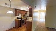 This single-floor residential space is designed with practicality and comfort in mind. The kitchen serves as a focal point, outfitted with rich, dark wood cabinetry that extends to the ceiling, providing generous storage throughout. Granite-look countertops offer durable workspace, while stainless steel appliances — including an electric range with overhead ventilation hood and a full-size refrigerator — lend a modern, polished feel. A peninsula-style counter adds additional prep space and subtly defines the kitchen's boundary. The flooring in the kitchen area consists of warm hardwood-style planks, adding an inviting contrast to the cabinetry. A textured ceiling runs throughout, contributing a classic residential character. Adjacent to the kitchen, a wide hallway carpeted in neutral tones leads toward additional rooms, hinting at a thoughtfully laid-out floor plan. The transition between the hard-surface kitchen and the soft-carpeted corridor creates a natural delineation of living zones. Overall, the space presents a well-organized, functional layout with cohesive finishes that balance warmth and utility throughout.