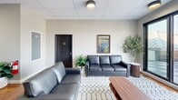 Located at 36 S Sixth St in Easton, PA, this contemporary space combines a well-appointed interior lounge with a striking rooftop terrace, offering a seamless blend of indoor comfort and outdoor living. The interior living room features warm hardwood flooring, a drop ceiling with recessed lighting, and clean neutral walls that create a bright and welcoming atmosphere. Sleek leather sofas are arranged around a patterned area rug, complemented by a bold dark-slatted accent wall that adds architectural depth and visual contrast. Lush potted greenery and curated artwork, including a local landscape painting, add warmth and character to the space. A corridor leads further into the building, hinting at additional amenities beyond. The rooftop terrace is a standout feature, finished with a checkered tile floor and enclosed by frameless glass railings that preserve unobstructed panoramic views of the Easton skyline and surrounding tree-lined neighborhoods. A circular wicker sectional seating arrangement anchors the outdoor lounge area, while an outdoor kitchen station offers functionality for entertaining. A wood-paneled exterior accent wall and mounted flat-screen television enhance the rooftop's resort-like ambiance, all beneath open skies that flood the space with natural light.