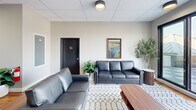 Located at 36 S Sixth St in Easton, PA, this contemporary space combines a well-appointed interior lounge with a striking rooftop terrace, offering a seamless blend of indoor comfort and outdoor living. The interior living room features warm hardwood flooring, a drop ceiling with recessed lighting, and clean neutral walls that create a bright and welcoming atmosphere. Sleek leather sofas are arranged around a patterned area rug, complemented by a bold dark-slatted accent wall that adds architectural depth and visual contrast. Lush potted greenery and curated artwork, including a local landscape painting, add warmth and character to the space. A corridor leads further into the building, hinting at additional amenities beyond. The rooftop terrace is a standout feature, finished with a checkered tile floor and enclosed by frameless glass railings that preserve unobstructed panoramic views of the Easton skyline and surrounding tree-lined neighborhoods. A circular wicker sectional seating arrangement anchors the outdoor lounge area, while an outdoor kitchen station offers functionality for entertaining. A wood-paneled exterior accent wall and mounted flat-screen television enhance the rooftop's resort-like ambiance, all beneath open skies that flood the space with natural light.