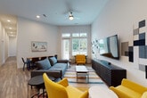 This two-story residence combines modern design with functional living across both levels. The open-concept living area serves as a central highlight, featuring wood-look flooring, elevated ceilings, and a ceiling fan. Large windows allow abundant natural light and outdoor views, while a striking feature wall with geometric acoustic panels in varying shades of gray frames a mounted flat-screen television. Vibrant yellow accent chairs and a colorful area rug add energy to the contemporary space, complemented by a separate dining area nearby.

The kitchen is outfitted with dark wood upper cabinetry, gray lower cabinets, white quartz countertops, a subway tile backsplash, and stainless steel appliances including a range, microwave, and dishwasher. A dedicated laundry room houses a stacked washer and dryer unit on wood-look flooring.

The bedrooms offer carpeted floors and generous windows that invite natural light and outdoor views. One room provides direct balcony access. The bathroom features gray cabinetry, a white quartz vanity, an undermount sink, and a large framed mirror.

A staircase with warm wood railings, white balusters, and carpeted treads connects both floors, adding a classic architectural touch to the overall contemporary aesthetic.