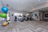 Located in Lawrenceville, GA, this well-appointed fitness center offers a comprehensive workout environment across a single floor. The space is finished with distinctive herringbone-patterned wood-look flooring that lends a polished, modern aesthetic throughout. Recessed lighting complements the abundant natural light streaming in through multiple large windows, providing bright and energizing views of the surrounding greenery and outdoor amenity areas.

The cardio zone is equipped with treadmills, elliptical trainers, and a stationary bike, arranged to maximize open floor space. A wall-mounted mirror enhances the sense of depth in this area. Strength training is well-supported with a multi-function cable machine and a dedicated leg press/extension station, offering a range of resistance-based workouts. A functional training corner rounds out the facility, featuring a wall-mounted stability ball rack stocked with multiple exercise balls in varying sizes, as well as a rack of resistance bands and workout mats for stretching and floor exercises.

The space includes double glass-panel exit doors that open directly to the outdoor area, reinforcing the connection to the property's exterior amenities. Neutral gray walls create a clean, calming backdrop, while the overall layout ensures ample room to move freely between equipment stations.